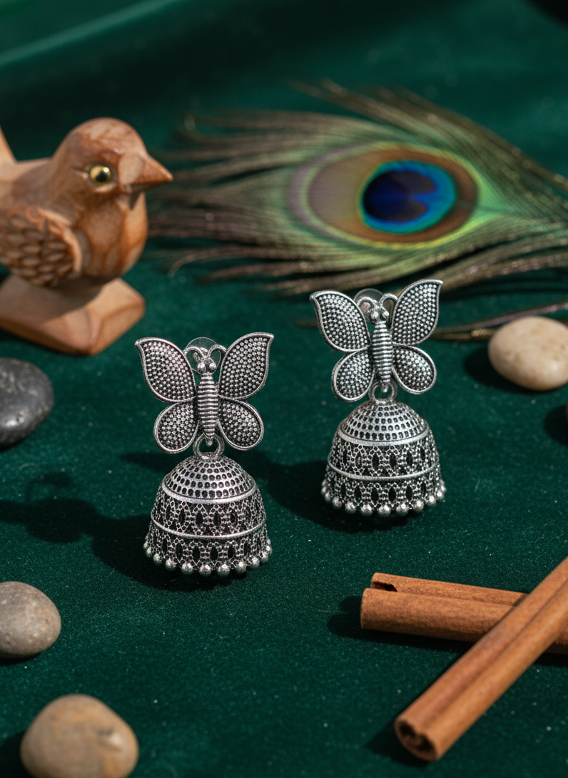 Silver butterfly jhumka earrings on a green surface with peacock feather and cinnamon sticks.