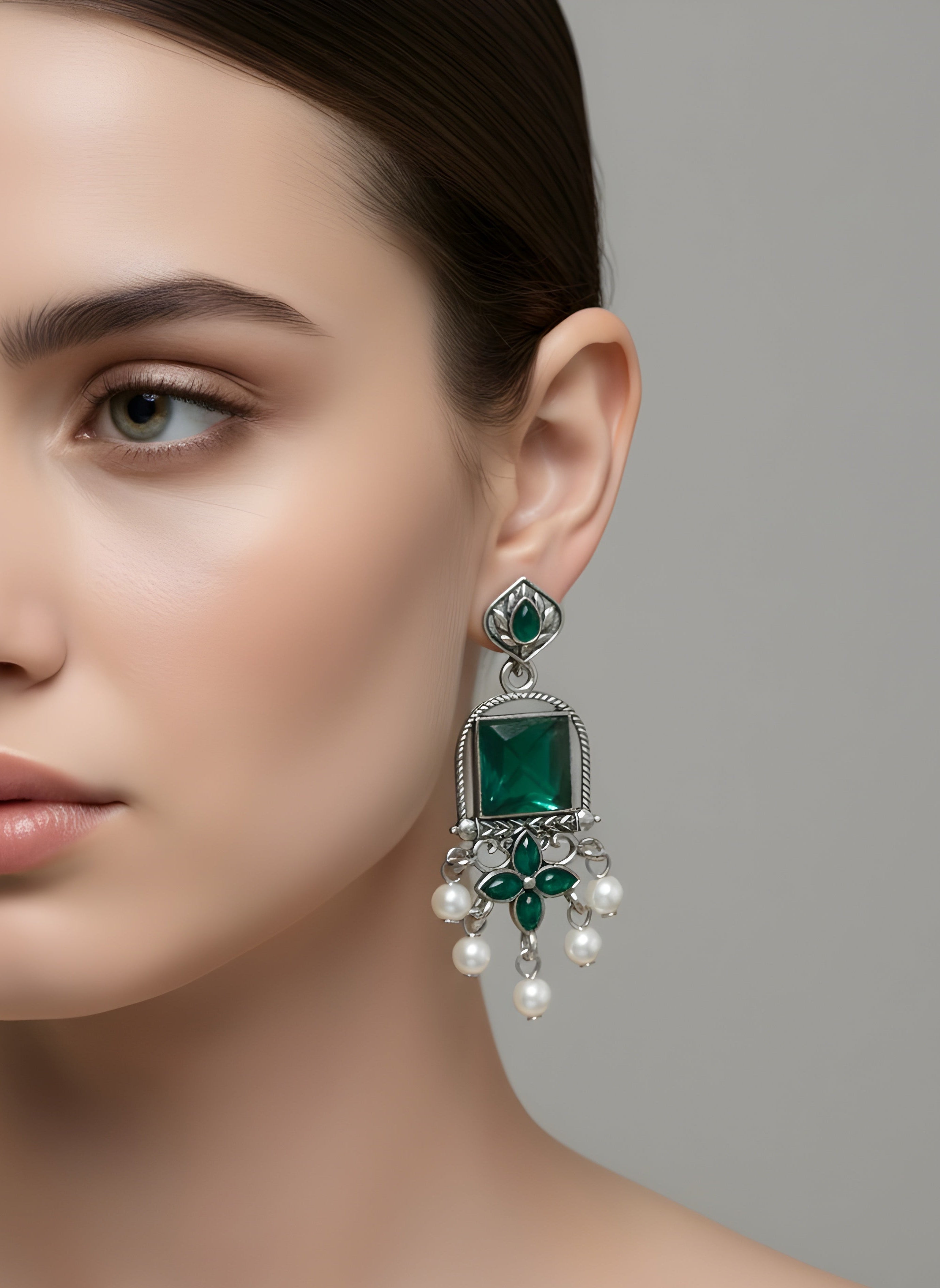 Close-up of a woman wearing emerald green and pearl earrings against a gray background