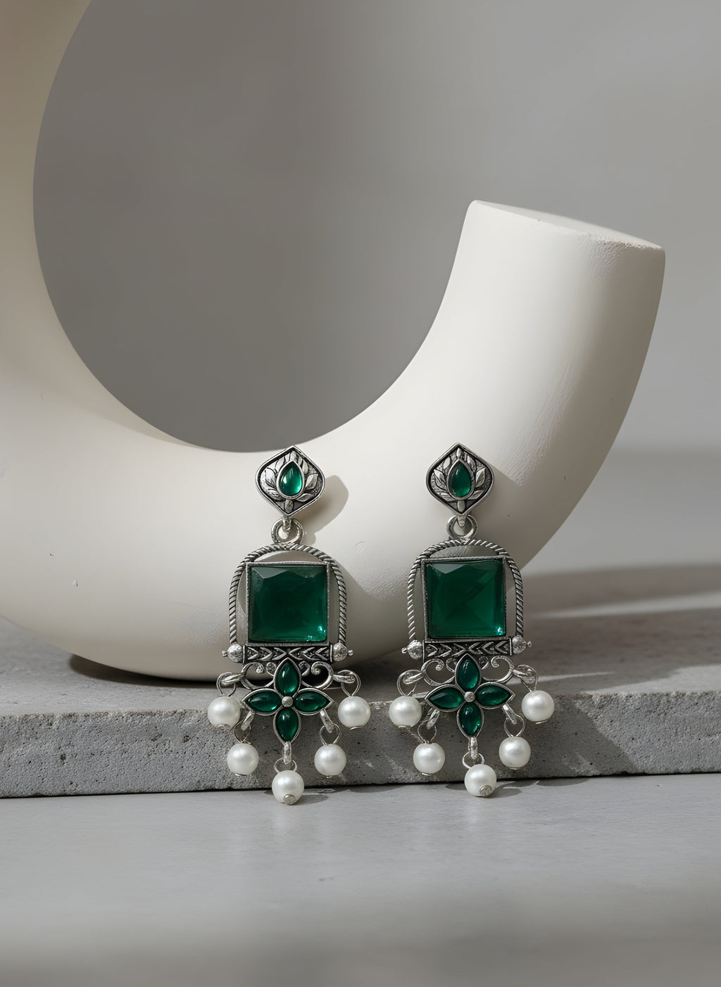 Green and silver earrings with pearl accents on a white ceramic holder.