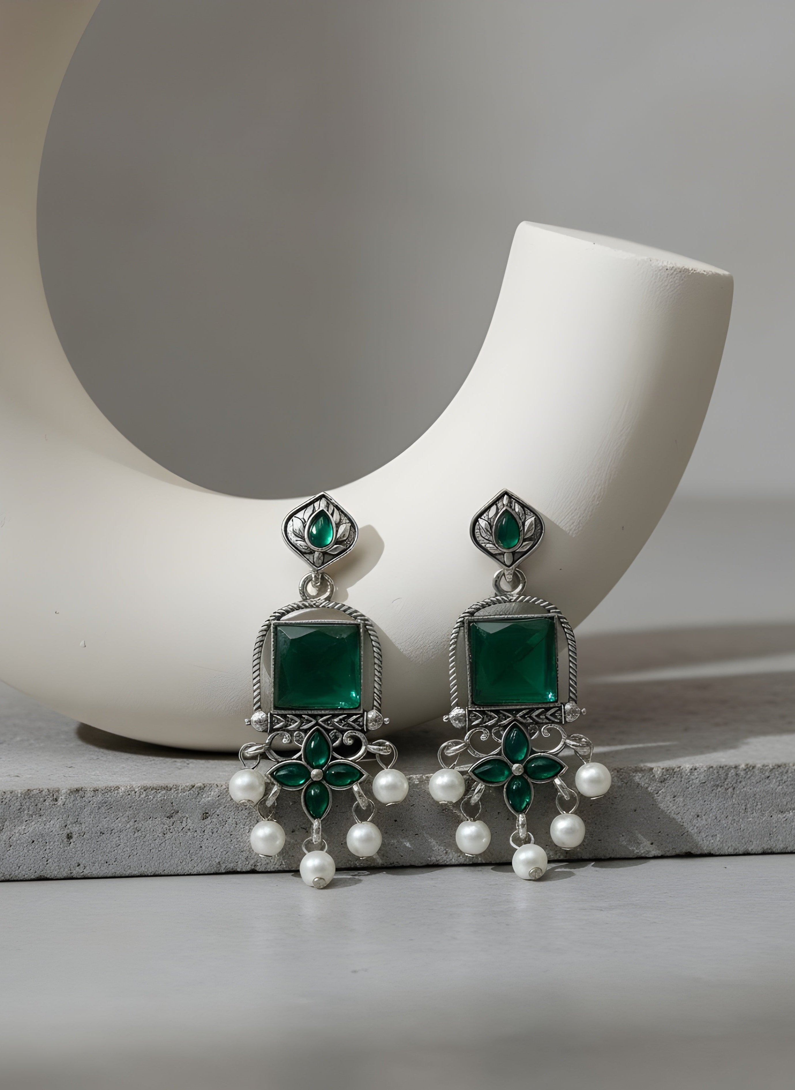Green and silver earrings with pearl accents on a white ceramic holder.