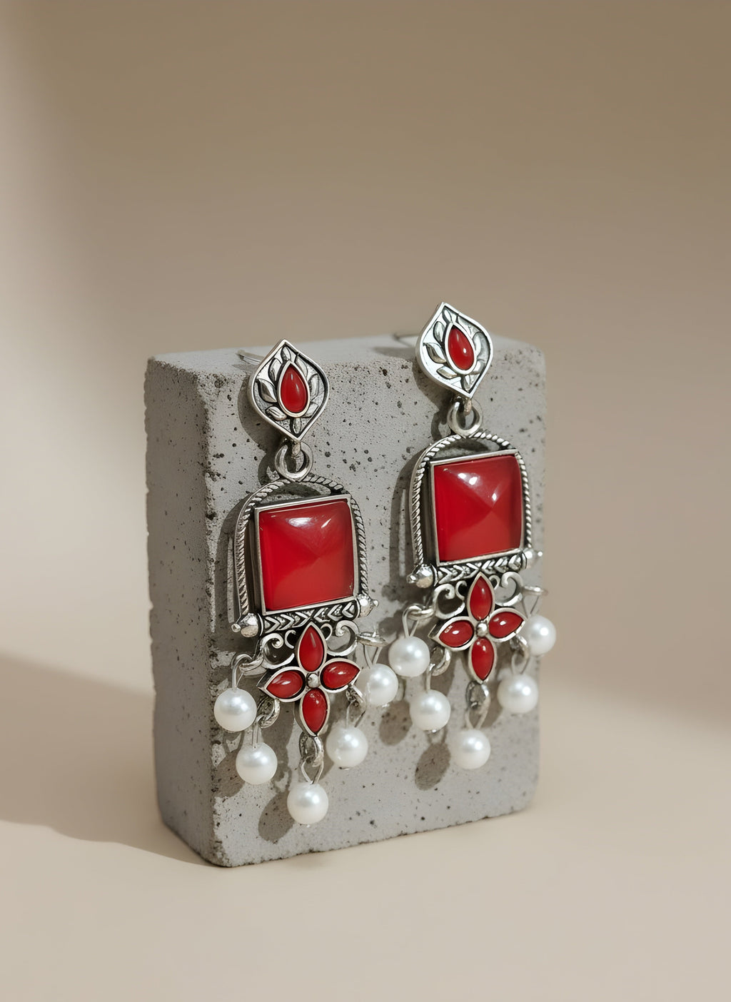 Pair of earrings with red stones and pearl-like elements on a concrete block against a beige background