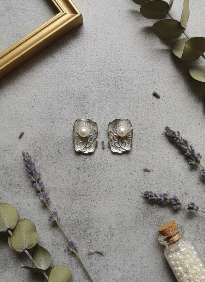 Silver earrings on a gray surface with eucalyptus and lavender, and a small bottle of pearls.