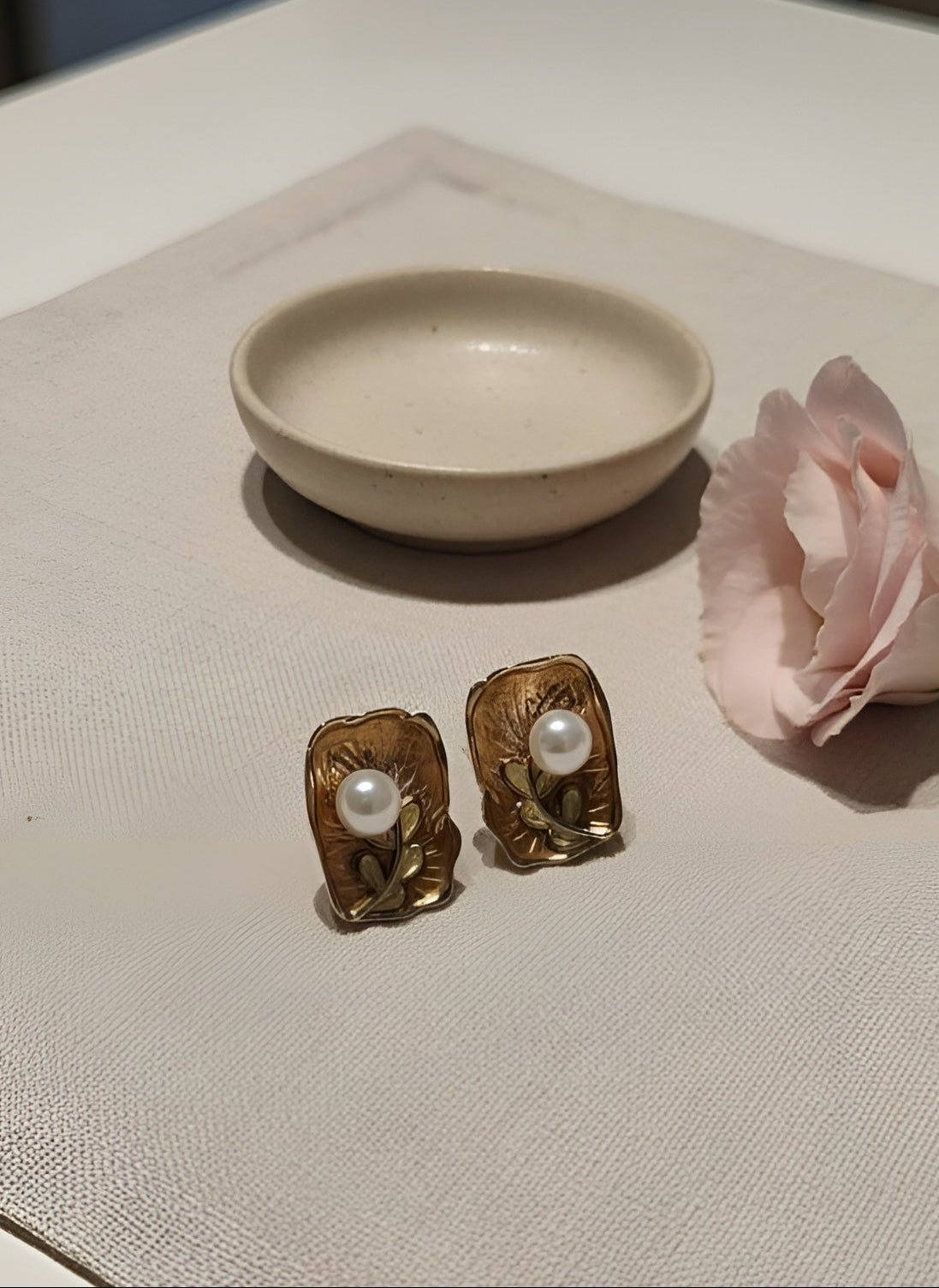 Gold earrings with pearls on a textured surface next to a pink flower and small dish.