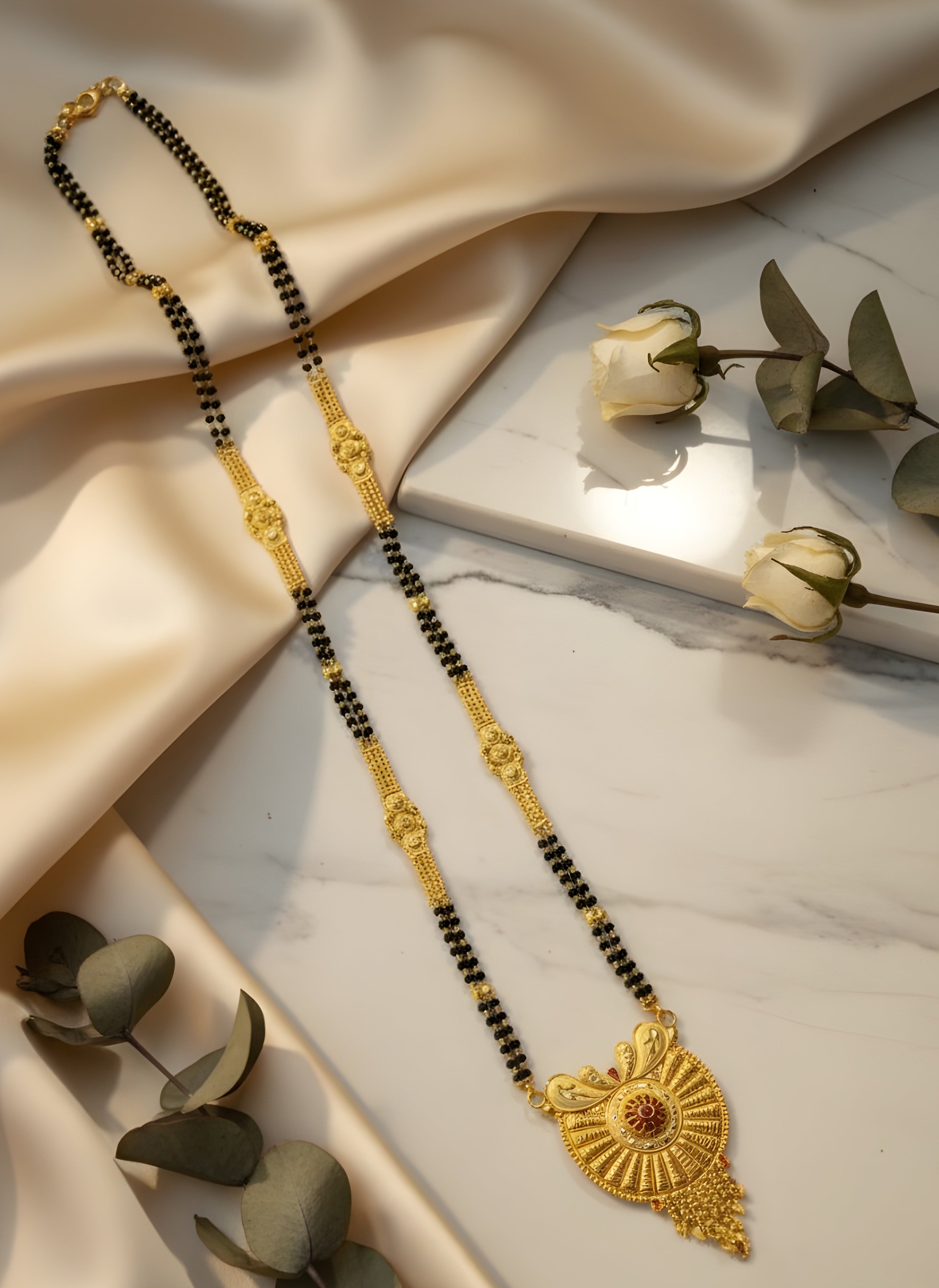 Gold and black beaded necklace on a marble surface with beige fabric and green leaves.
